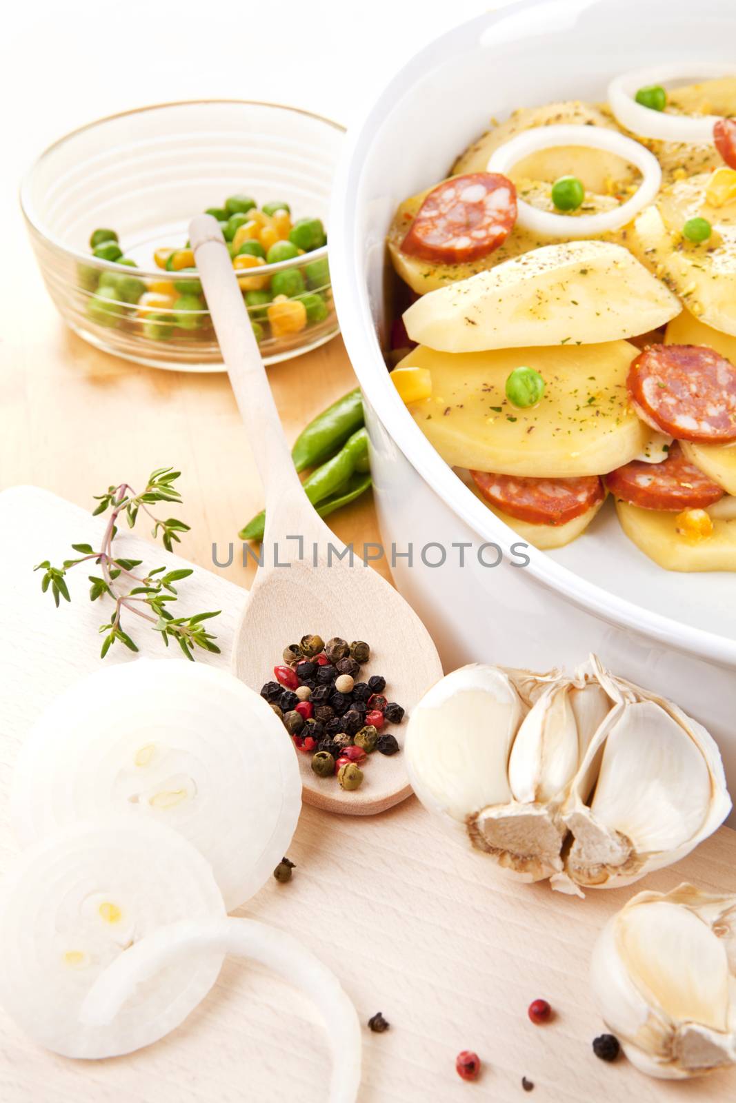 Fresh potatoes slices with fresh vegetables and sausage prepared for baking in bowl on wooden table.