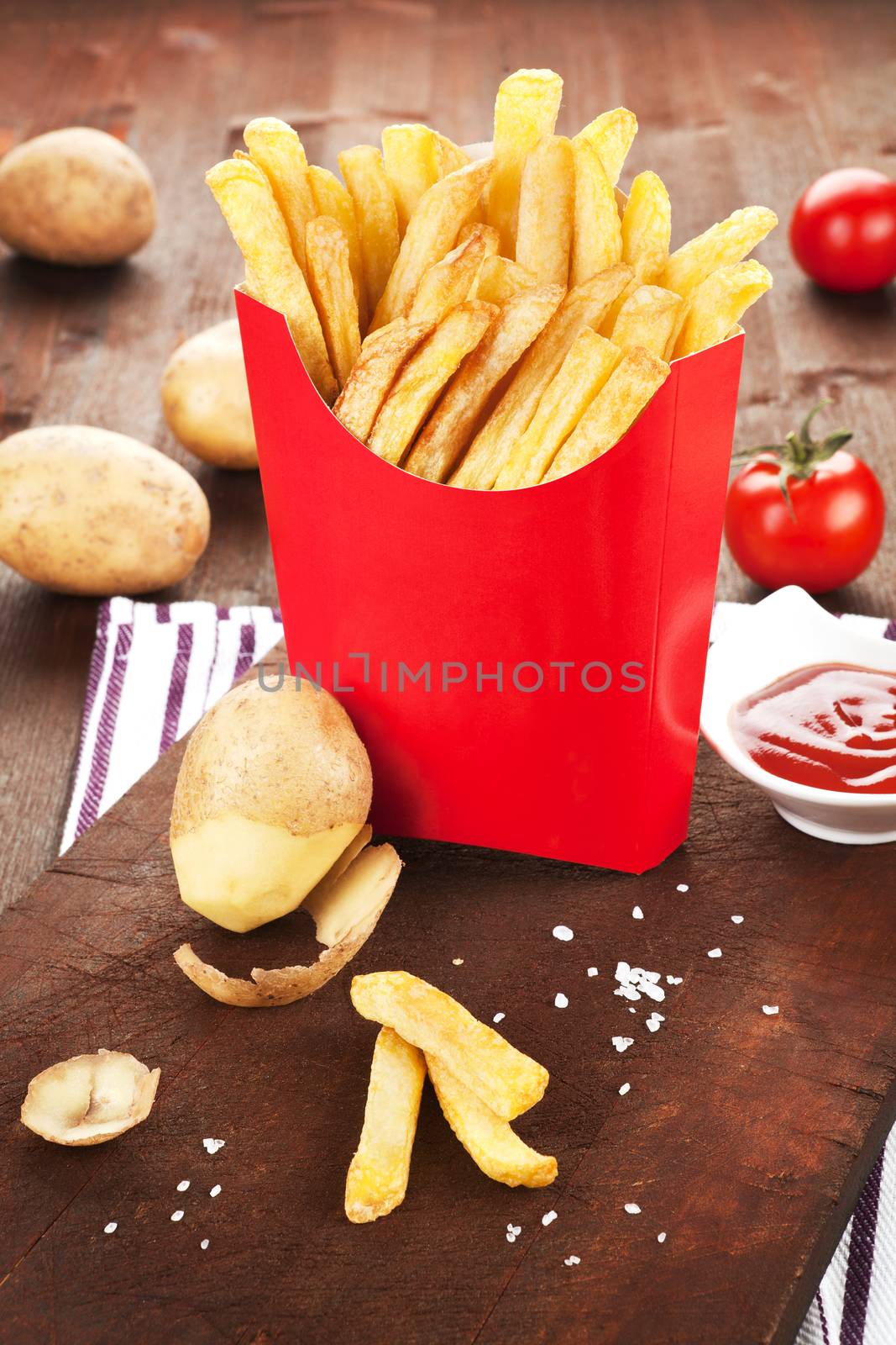 French fries in fast food paper bag. Potatoes, tomatoes and ketchup in background. Country style.