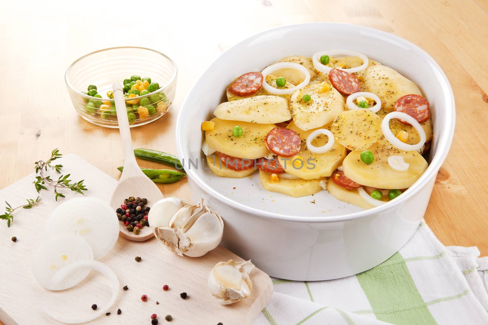 Fresh potatoes slices with fresh vegetables and sausage prepared for baking in bowl on wooden table.