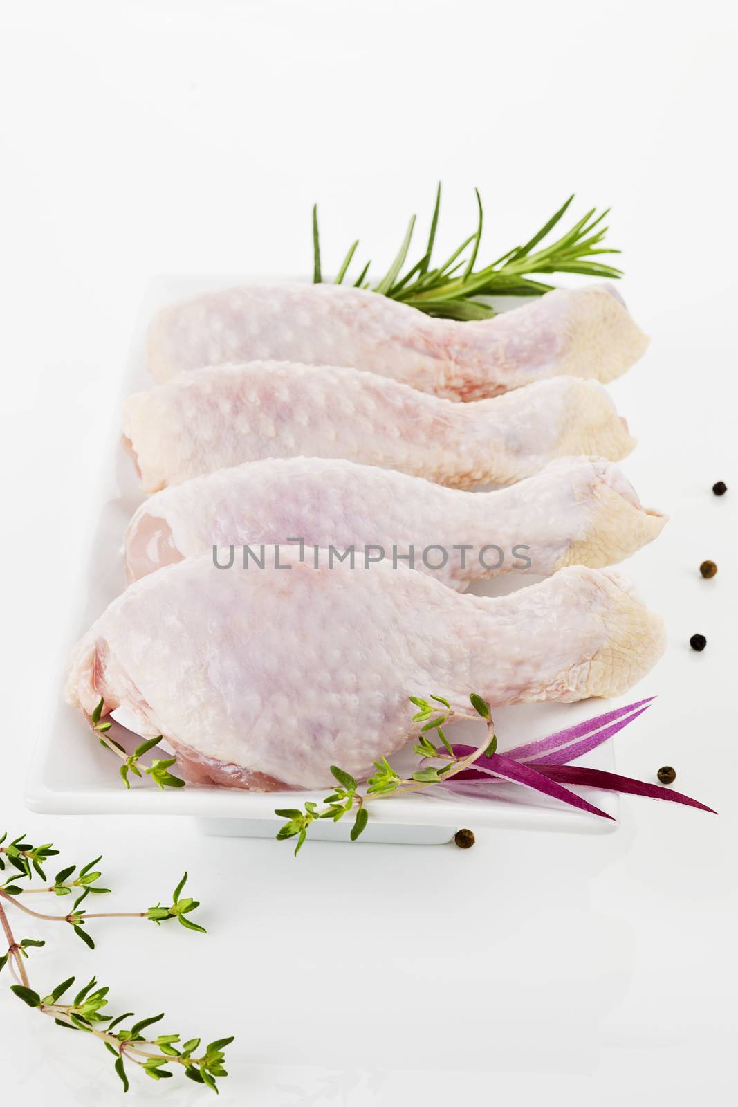 Four uncooked chicken legs on white plate decorated with rosemary and pepper corns.