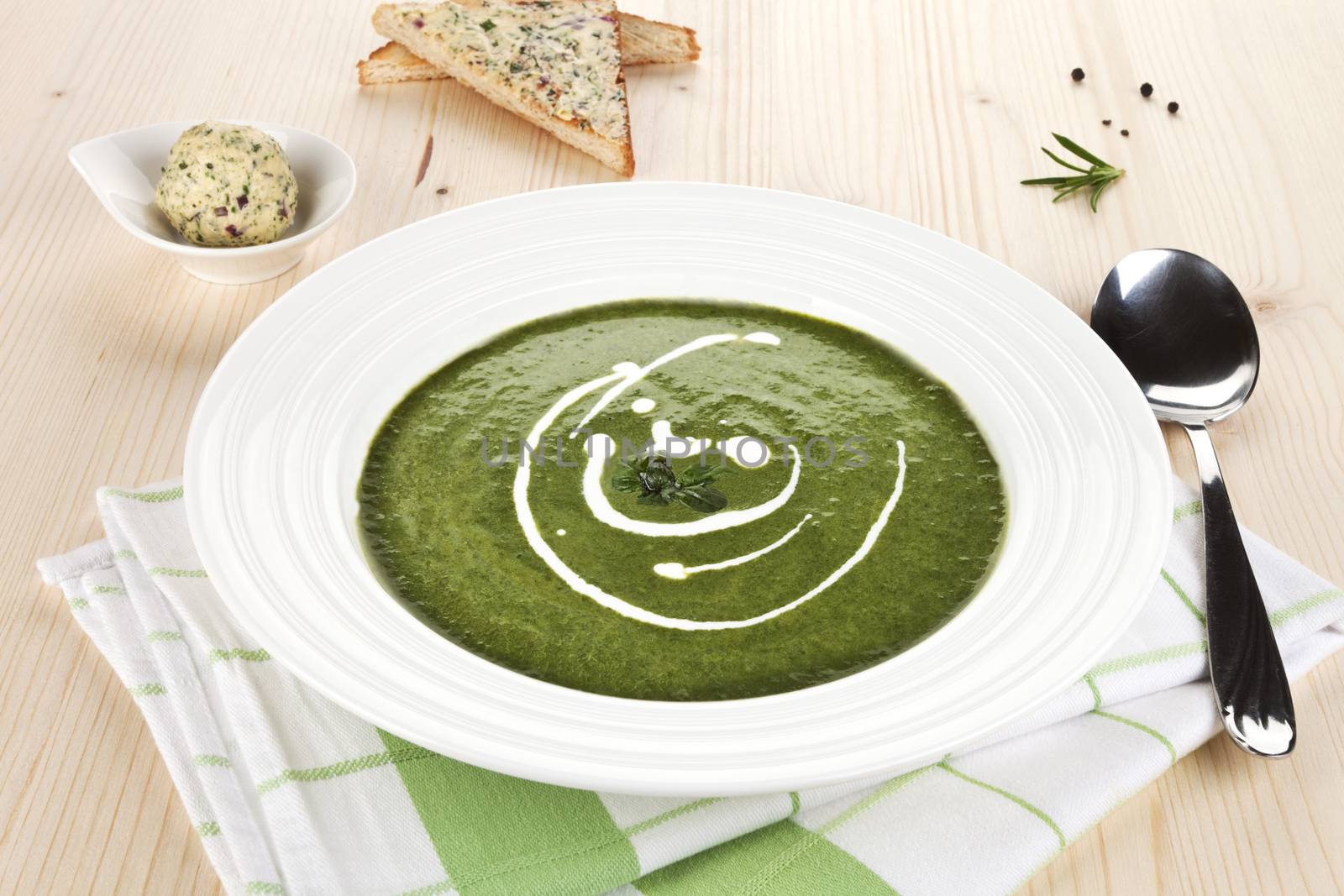 Spinach soup in white plate on wooden table, toast bread and herbal butter in bowl in background.