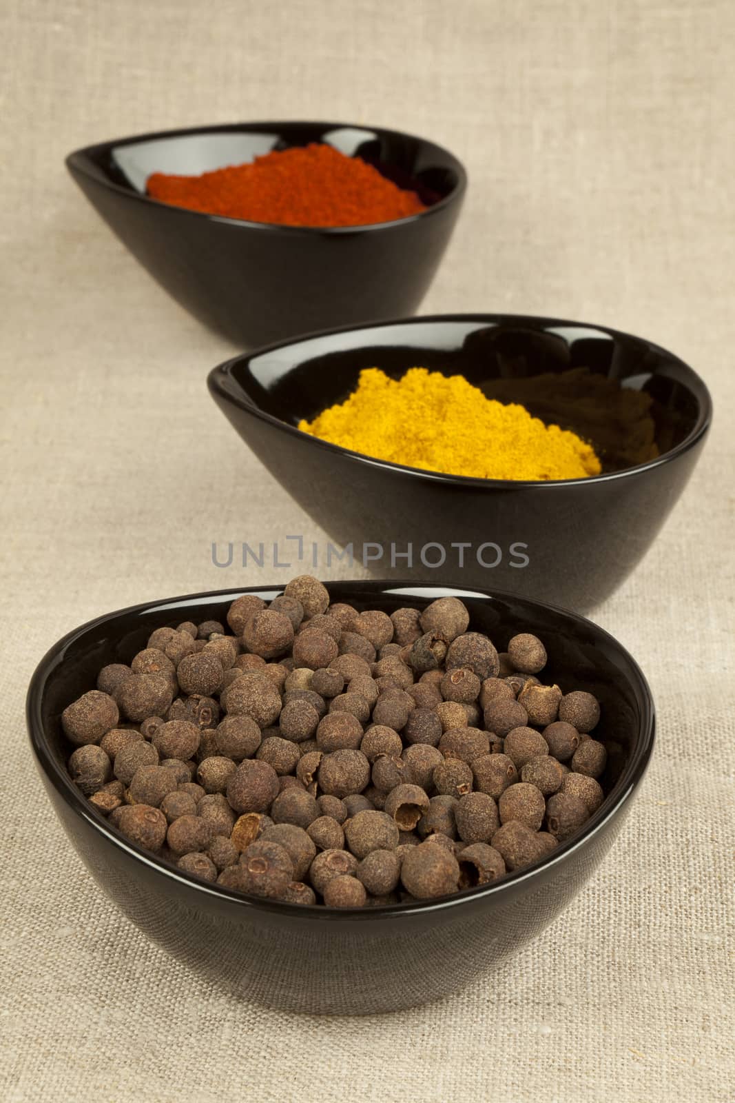 Three black bowls with different spices on brown natural textile background. Pepper corns, paprika and curry.