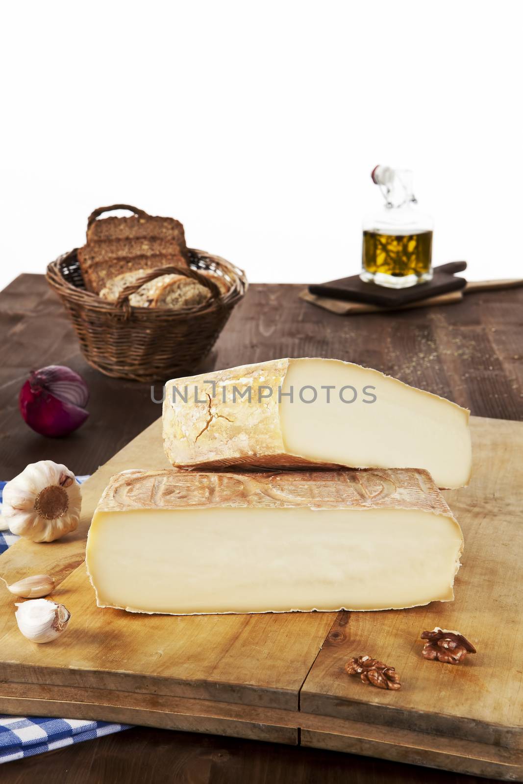 Big cheese pieces arranged on wooden board with garlic, onion, olive oil and bread in basket.