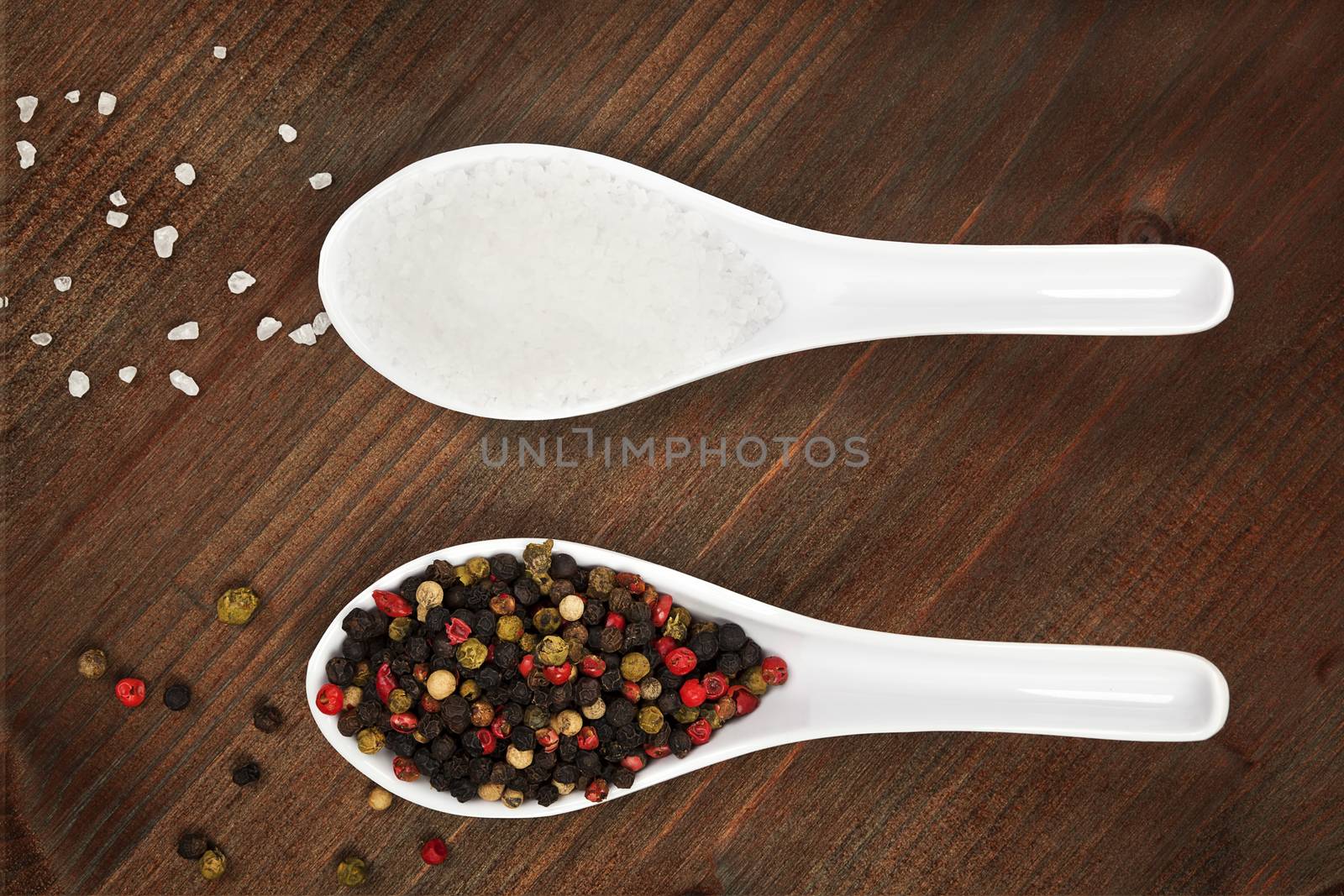 Salt crystal and pepper corns on white spoon on dark wooden background.