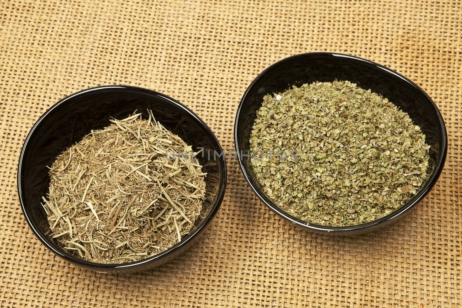 Rosemary and marjoram spices in black round bowls isolated on brown background.