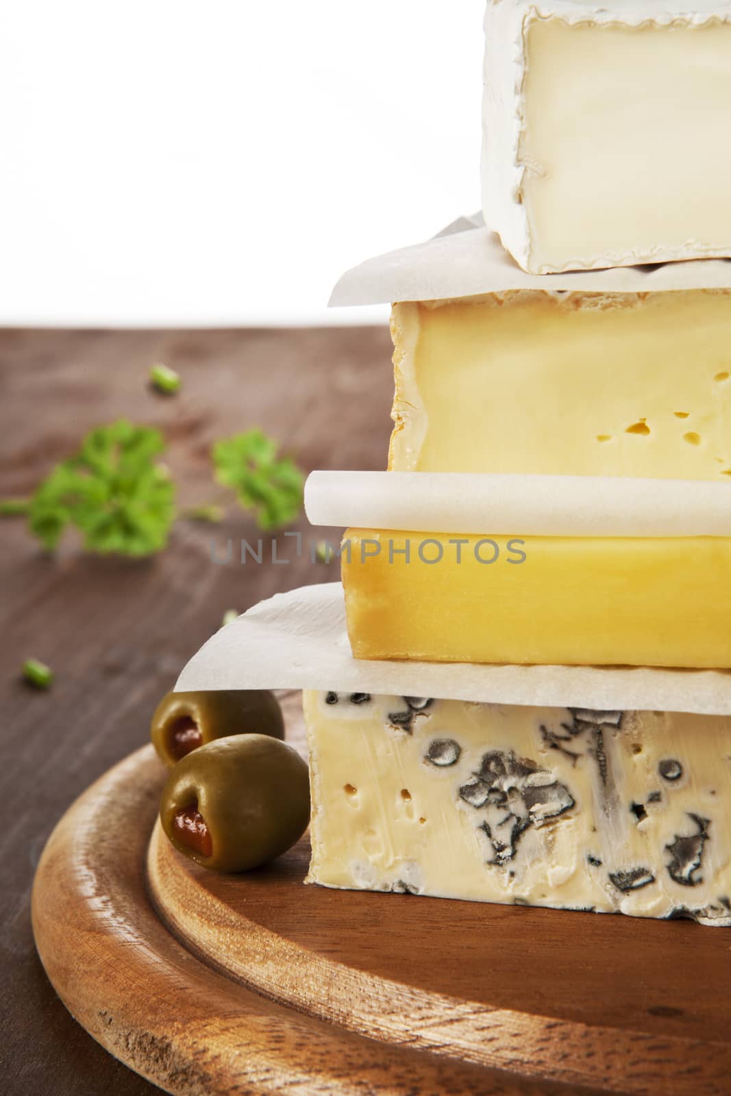 Various cheese pieces arranged on each other on brown wooden natural background with olives and parsley in background. Cheese background.