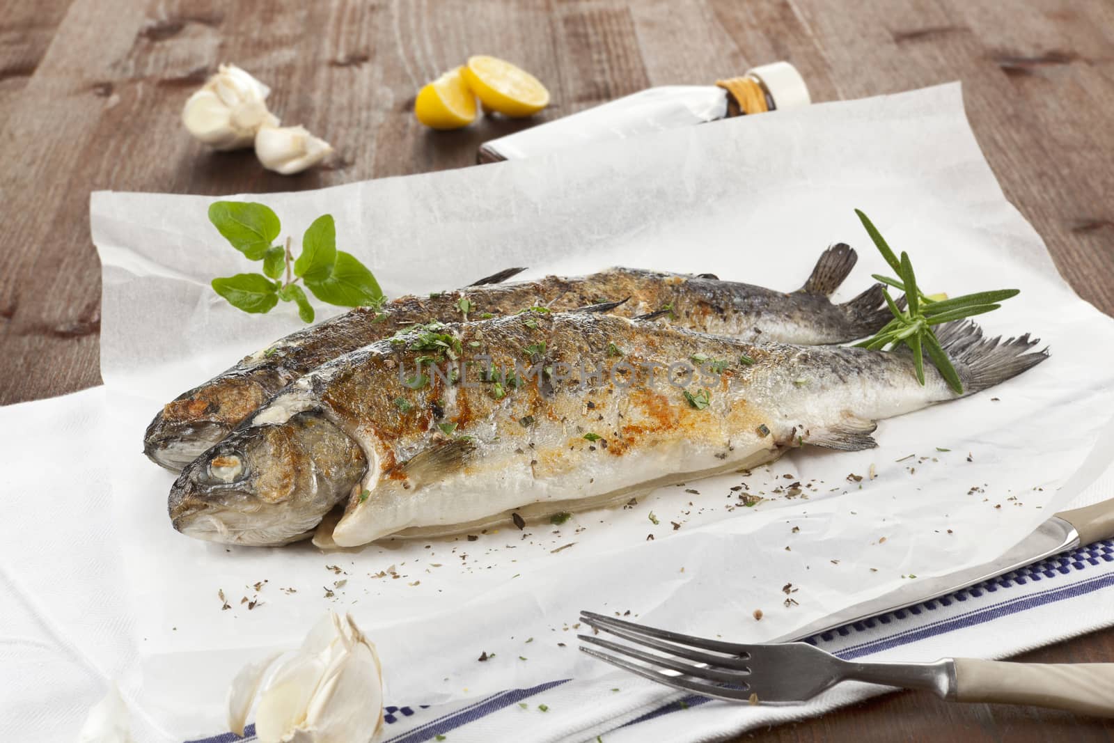 Two grilled trouts with lemon pieces, fresh herbs and garlic on white baking paper on kitchen towel on wooden table.