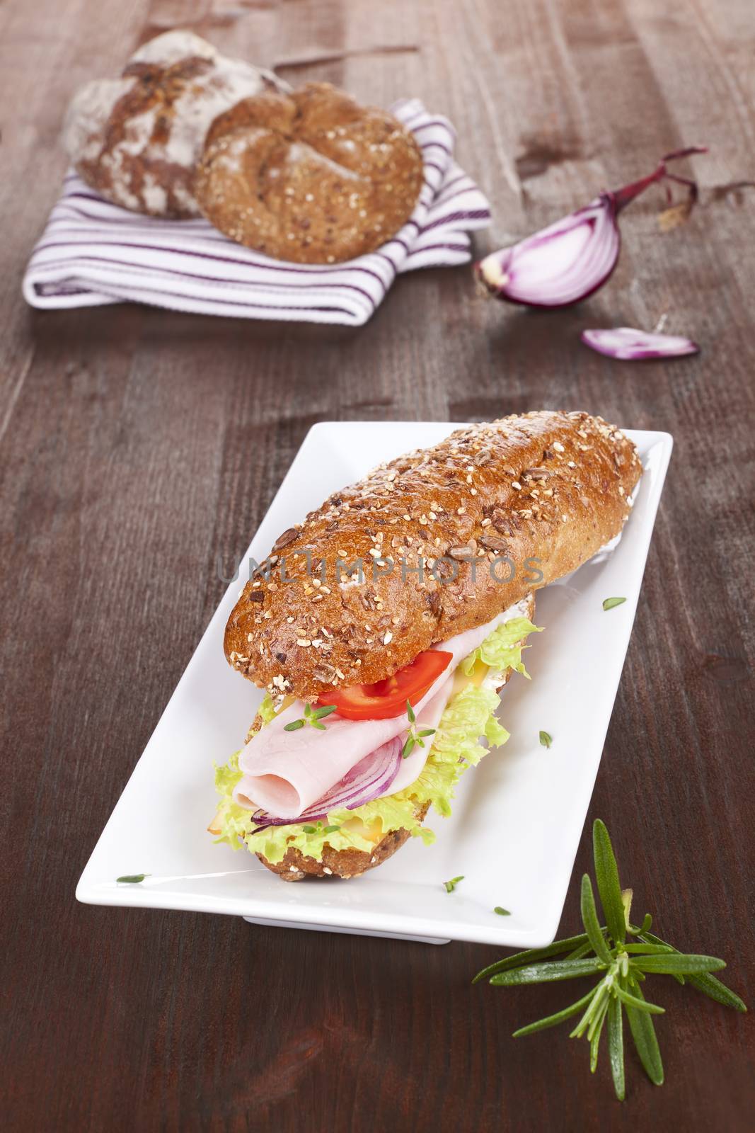 Wholegrain baguette with ham and fresh vegetables, decorated with onion and rosemary on brown wooden background.