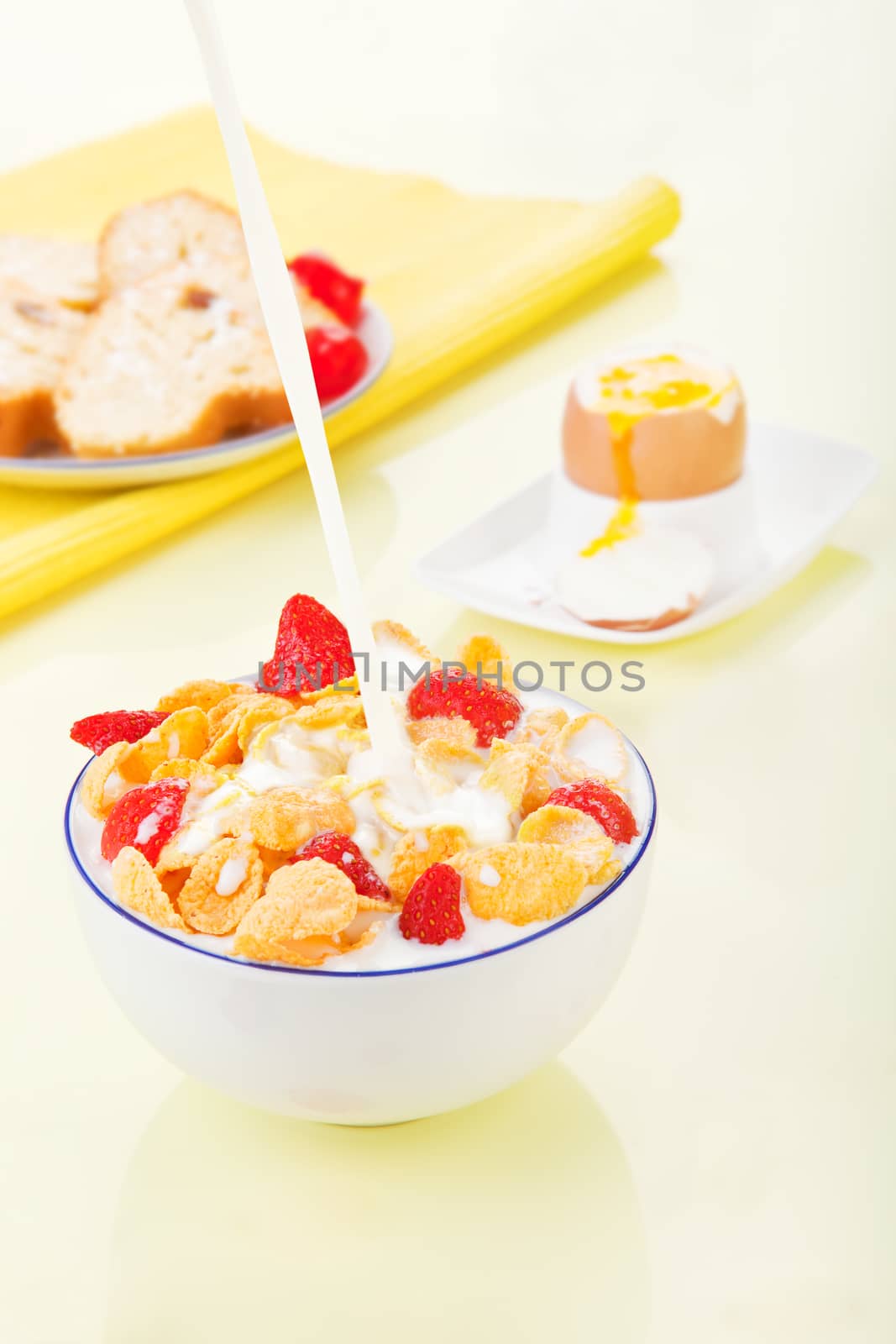 Pouring milk into a bowl with corn flakes and strawberries. Delicious breakfast. Egg and pastry in background.