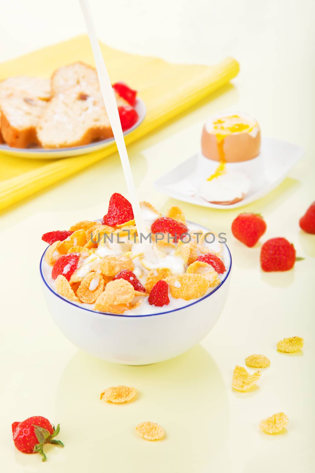 Pouring milk into bowl with corn flakes with strawberries. Delicious breakfast. Corn flakes with strawberries in bowl, bread and egg in background.