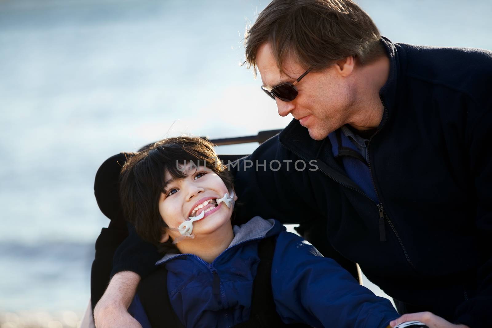 Father sitting with disabled son in wheelchair by lake shore