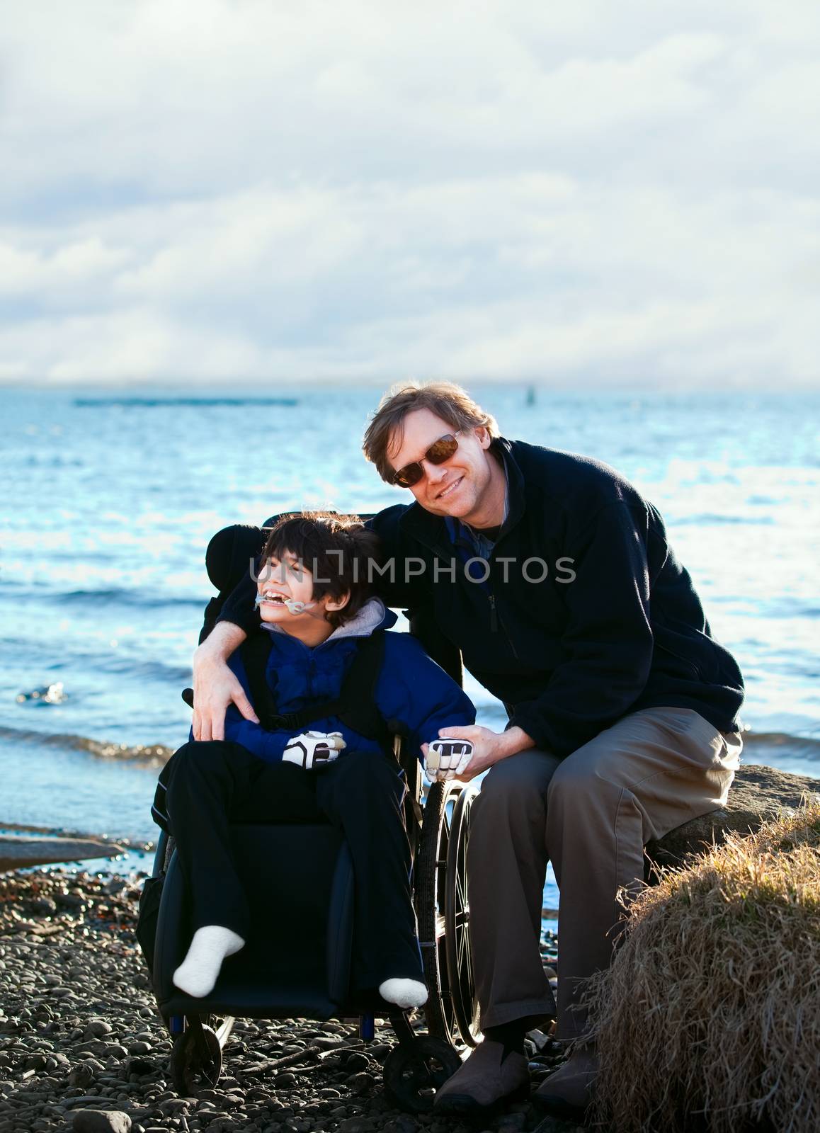 Father sitting with disabled son in wheelchair by lake shore