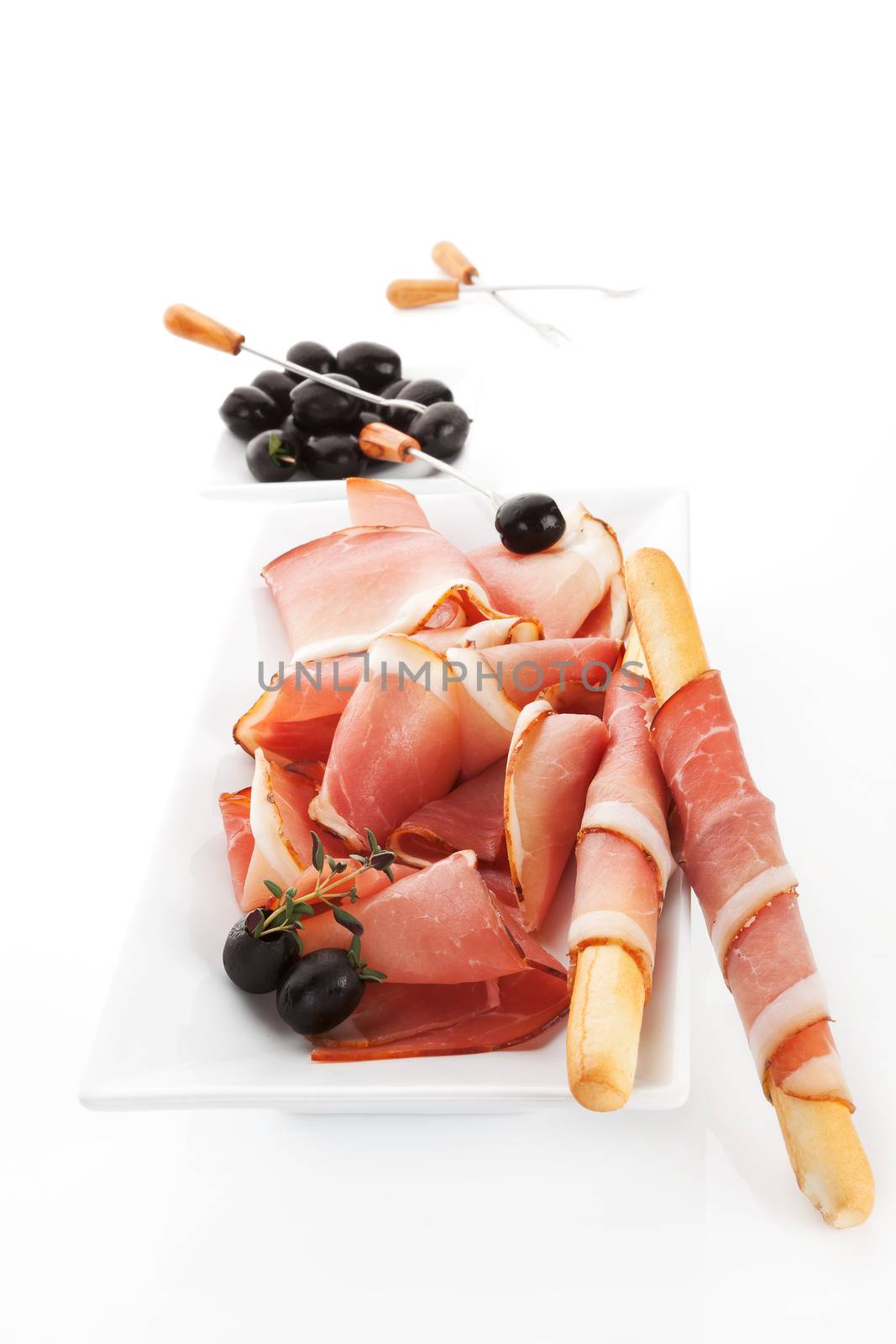 Delicious prosciutto slices with grissini breadsticks, black olives and thyme on white tray isolated on white. Traditional culinary meat background.