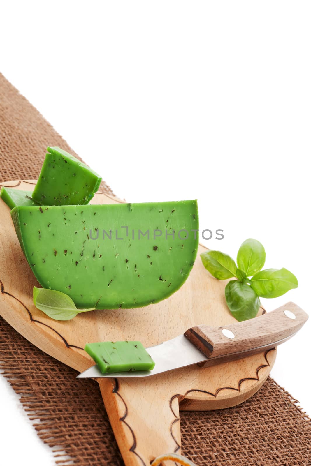 Delicious green pesto gouda on round decorated cutting board with knife on brown textile with copy space for your text. Culinary eating concept.