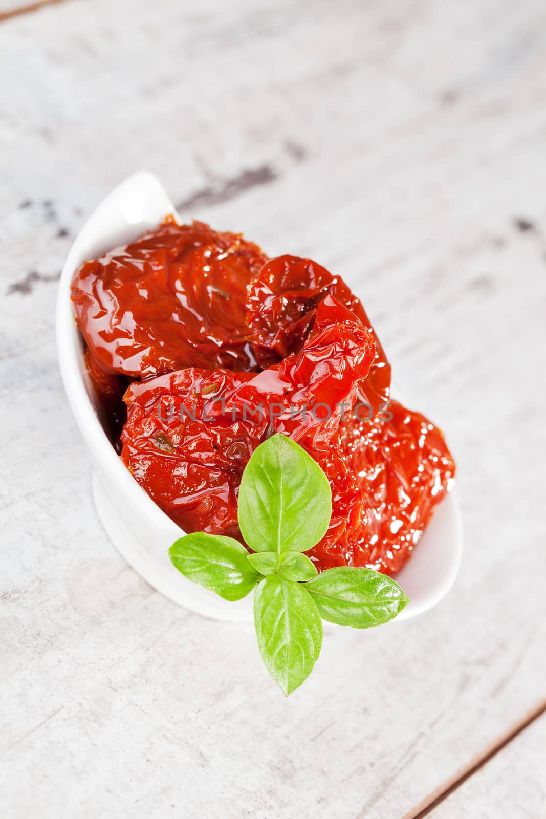 Delicious dried tomatoes with fresh basil leaves in bowl on white textured wooden background. Culinary eating