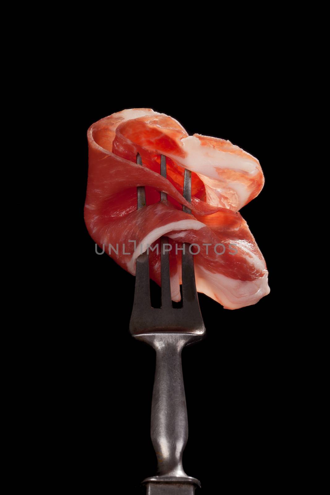 Delicious parma ham slice on old silver fork isolated on black background. Culinary traditional italian parma ham concept. Prosciutto.