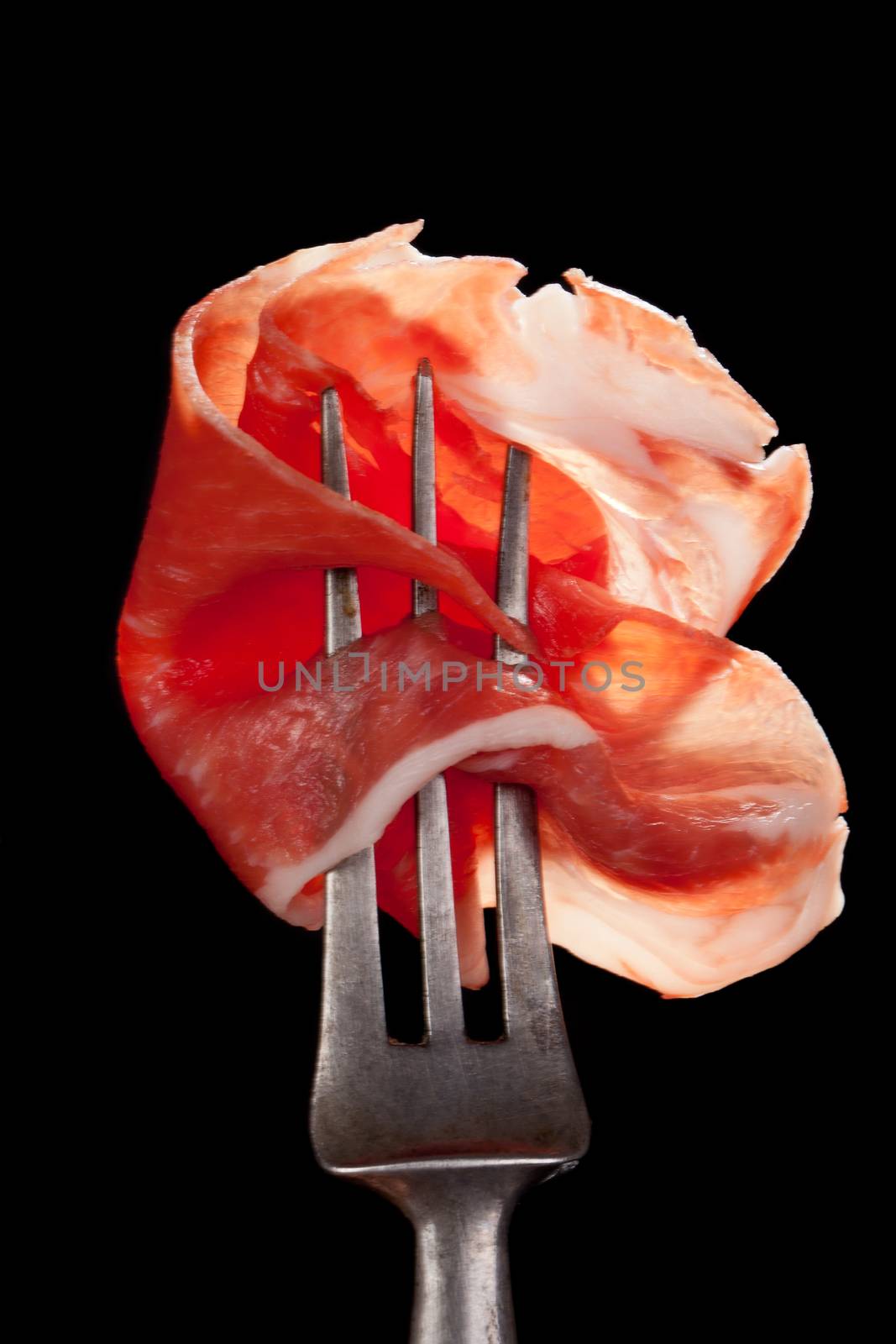 Delicious parma ham on old silver fork detail isolated on black background. Culinary traditional meat eating concept.