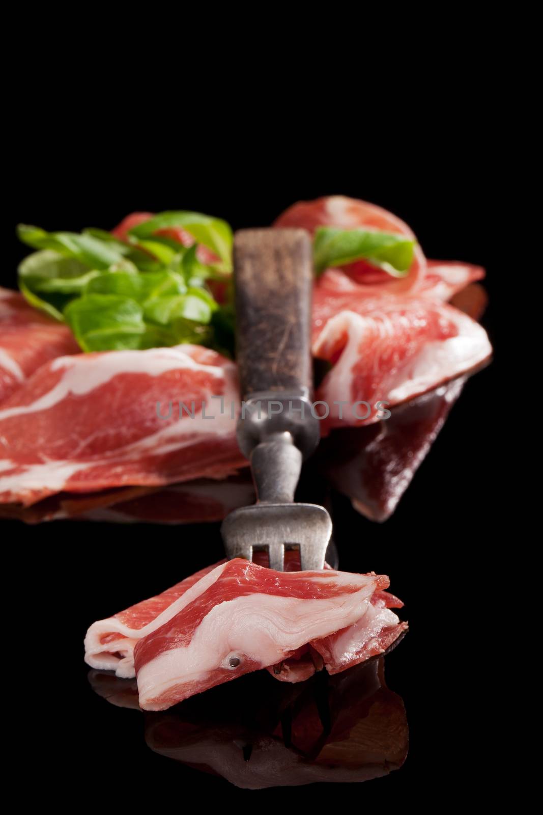 Prosciutto slices with fresh salad and old fork isolated on black. Rustic style country ham background.