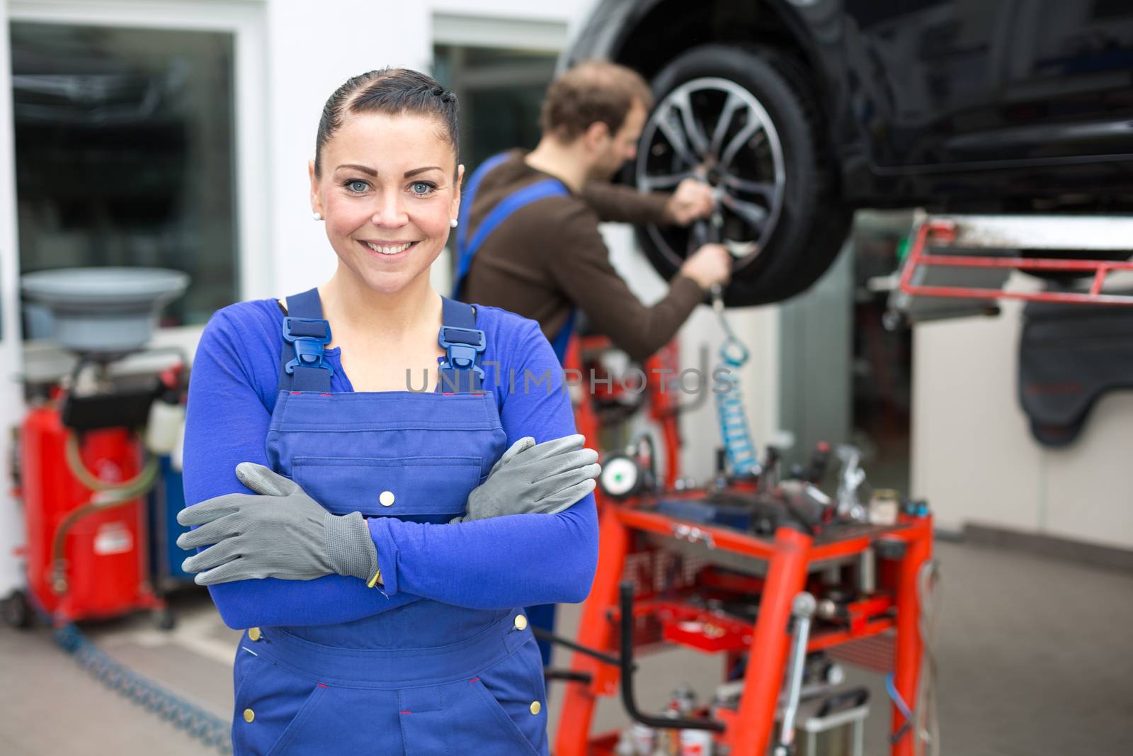 Female mechanic standing in workshop, another mechanic changes a wheel at a car on a hydraulic lift