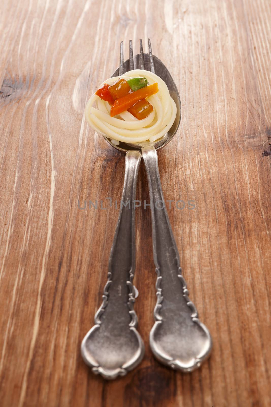 Culinary pasta eating. Pasta with colorful vegetable on silver spoon with fork isolated on brown wooden background.