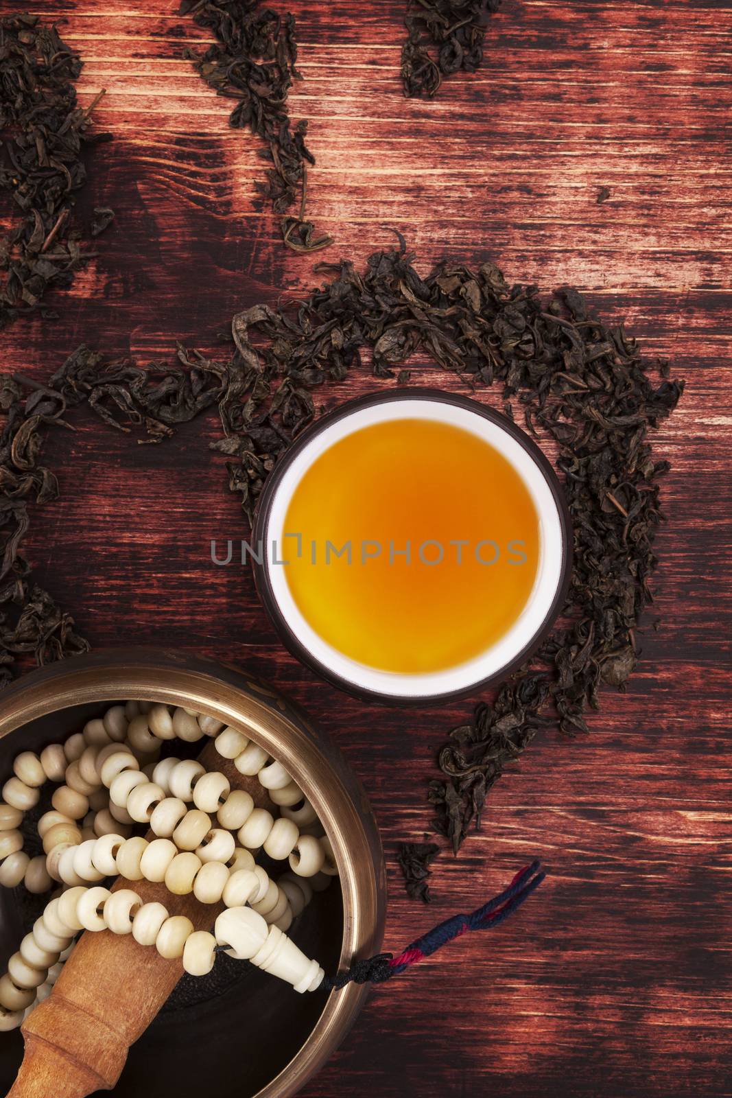 Traditional tea ritual. Tea in cup, dry tea crop forming the ohm symbol, tibetan singing bowl with buddhist necklace on textured brown wooden background, top view. Harmony, buddhism and spirituality.