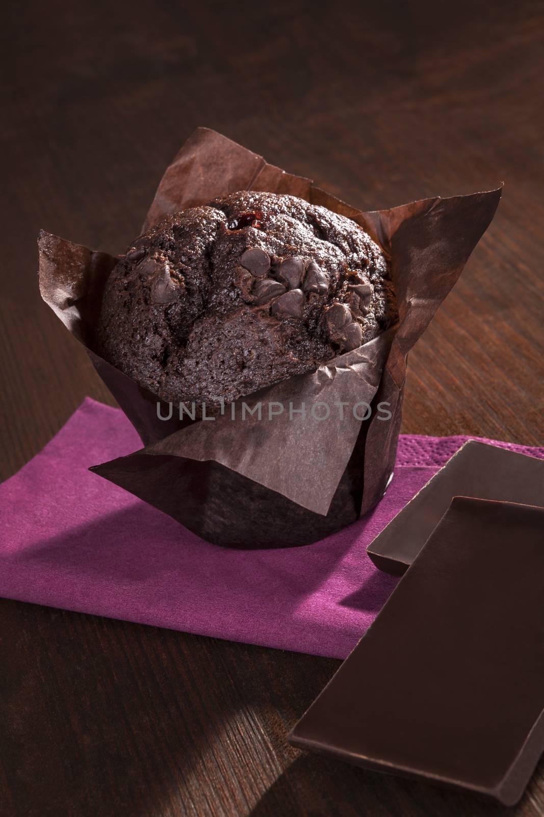 Delicious chocolate muffin with chocolate bar on purple napkin on wooden background. Dark brown culinary sweets.