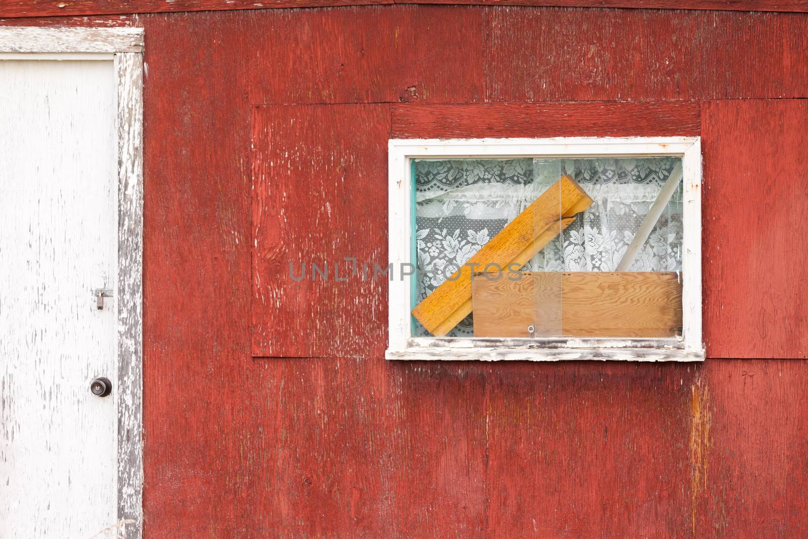 Exterior window on weathered wooden cabin with curtains and blocks of wood behind the glass next to shabby white painted door