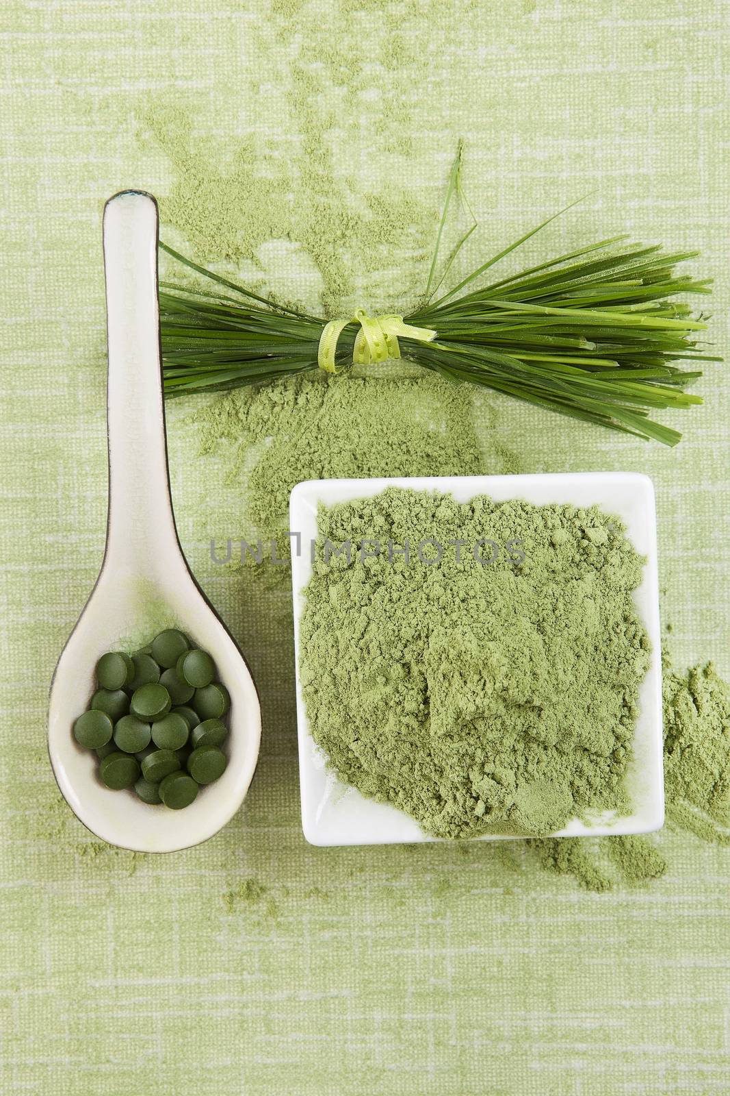 Green superfood. Green pills, green ground powder and wheat grass blades on green background. Chlorella, spirulina, wheat grass and barley grass.