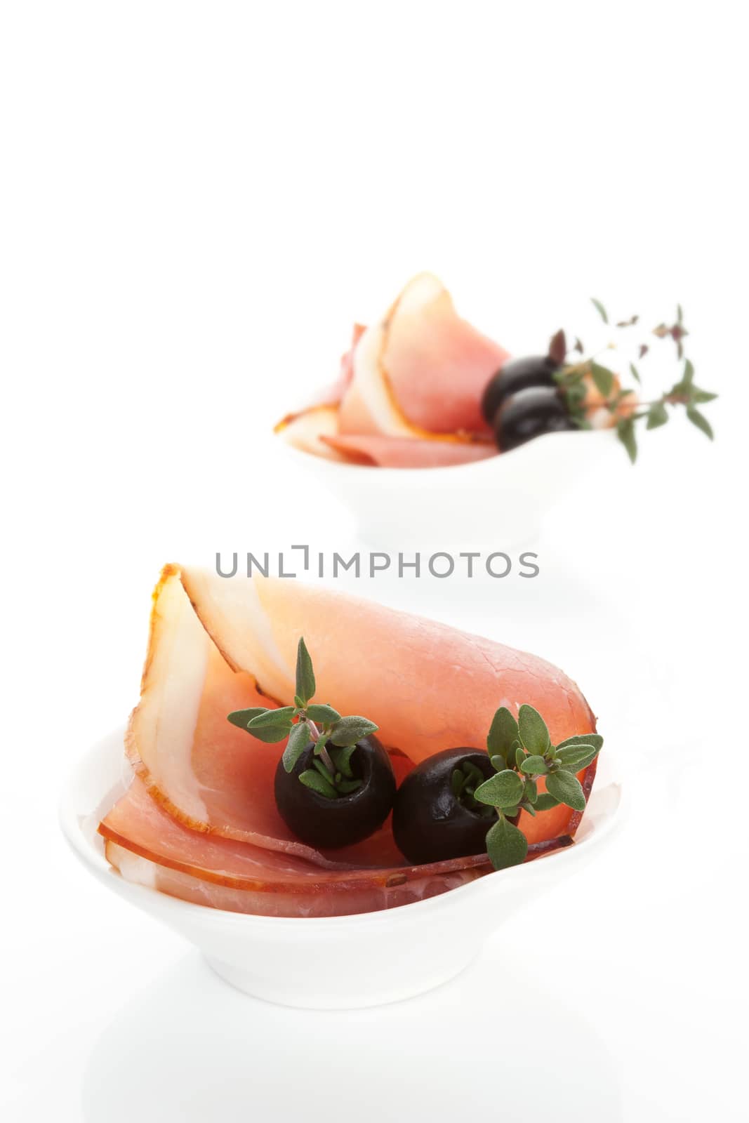 Prosciutto slices with black olives and fresh herbs in white bowl isolated on white background. Luxurious culinary canape.