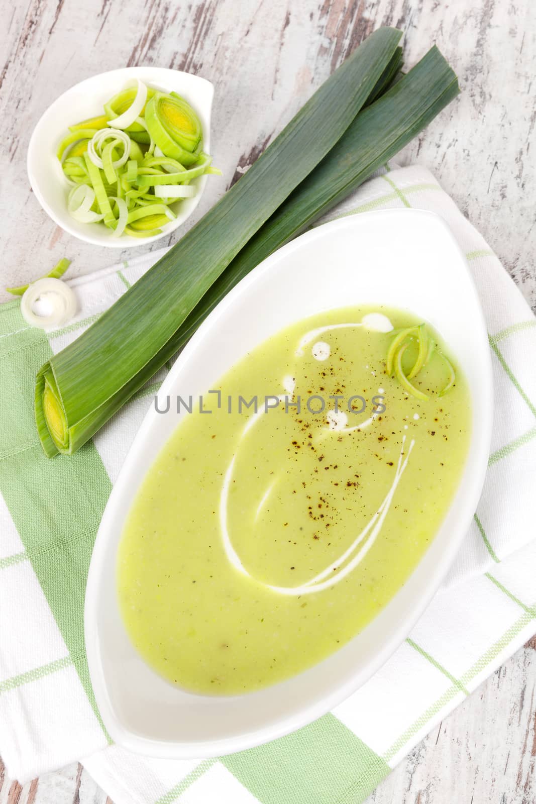 Delicious leek soup in white bowl with fresh and cut leek on white wooden background. Culinary healthy eating, top view.