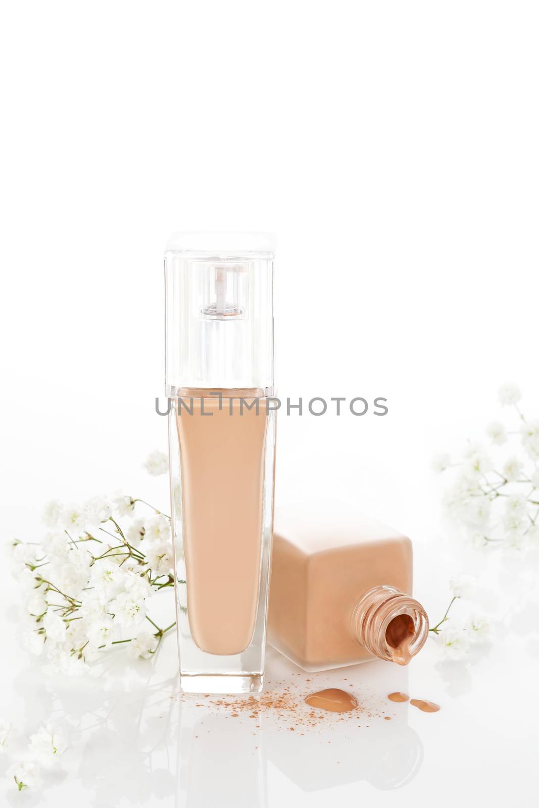 Beige liquid makeup dropping from luxurious glass container with white blossom isolated on white background with reflection. Glamour feminine cosmetics concept.