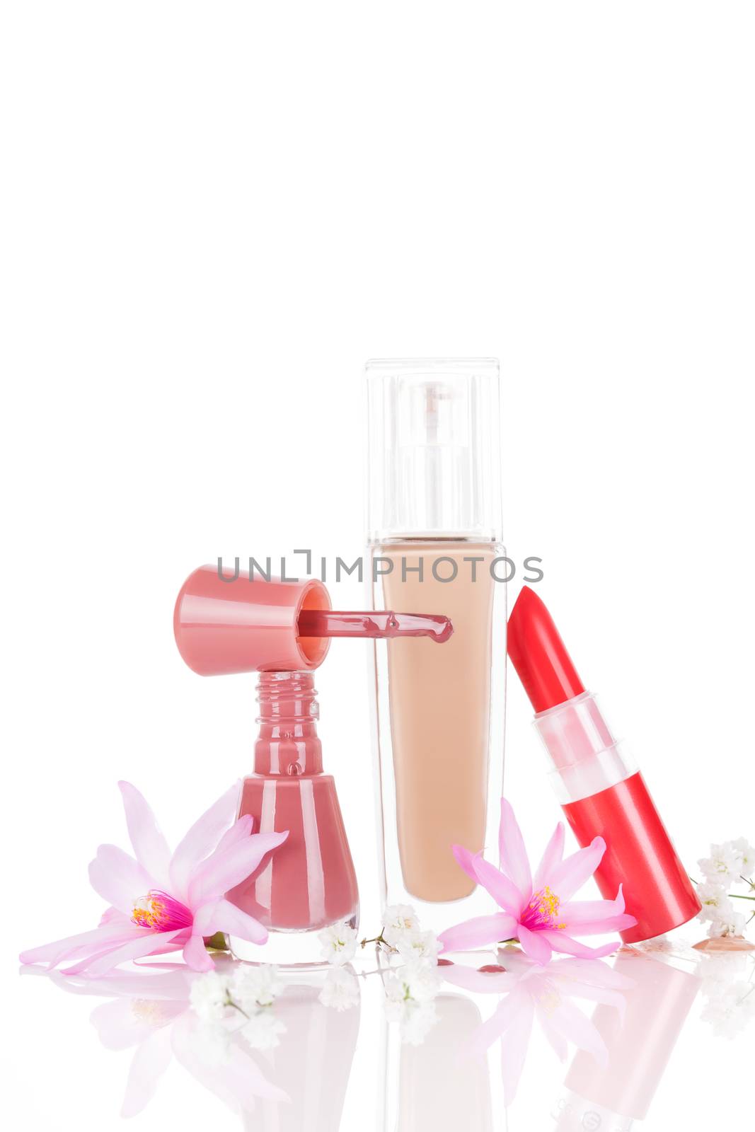 Luxurious makeup and cosmetic still life. Nail polish, red lipstick and beige foundation with pink and white flowers isolated on white background with reflection. Glamour beauty concept.