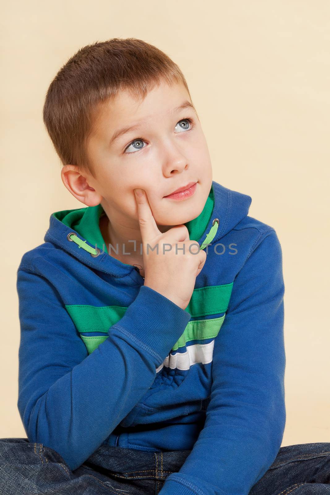 Young cute boy in casual blue clothing sitting, looking up, touching his chin and thinking isolated.