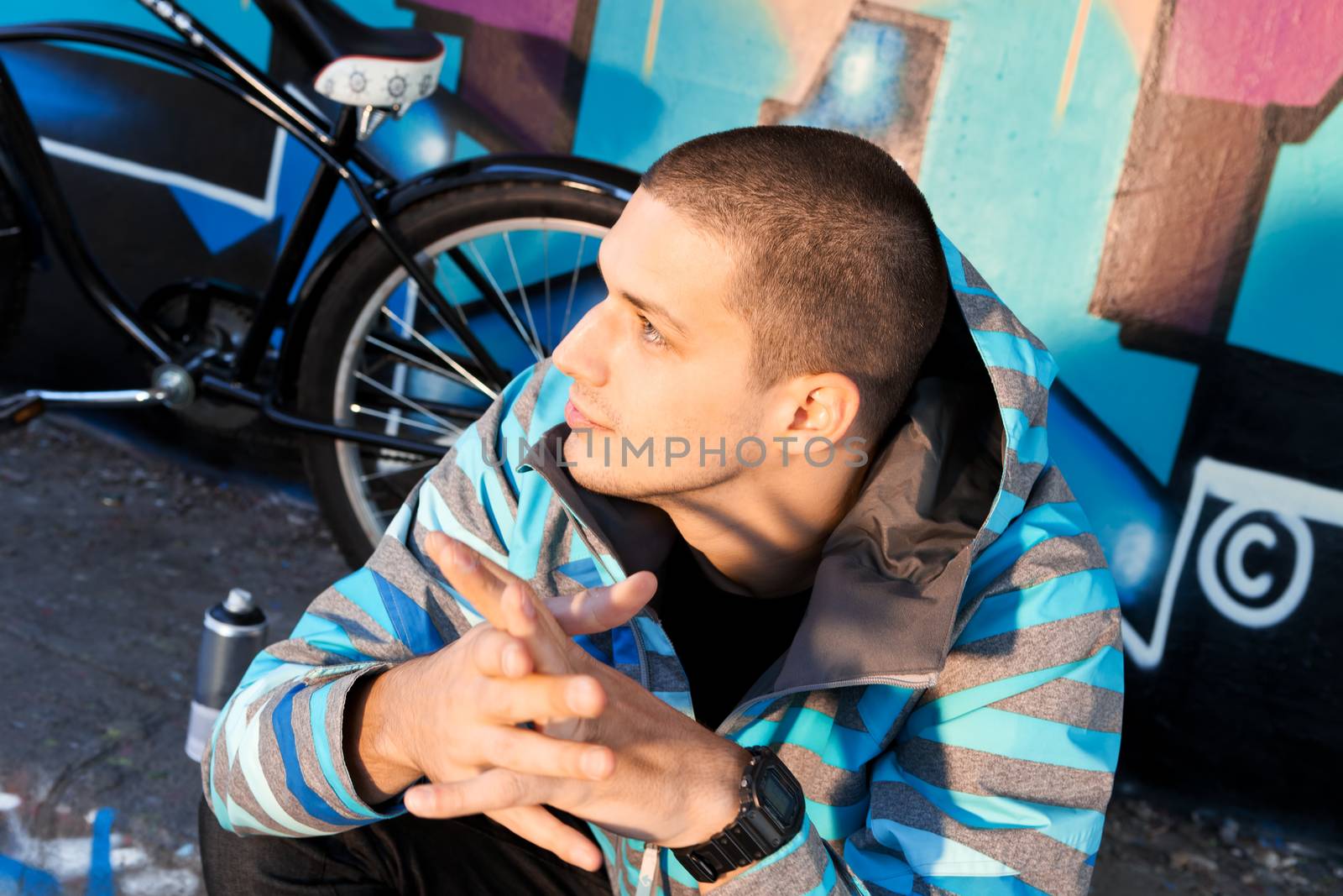 Young male graffiti artist sitting in front of finished piece on wall with city cruiser bike leaning next to him. Urban culture.