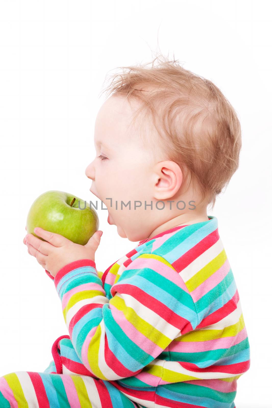 Small cute baby girl in colorful clothing holding and eating green apple isolated on white background. First teeth concept.