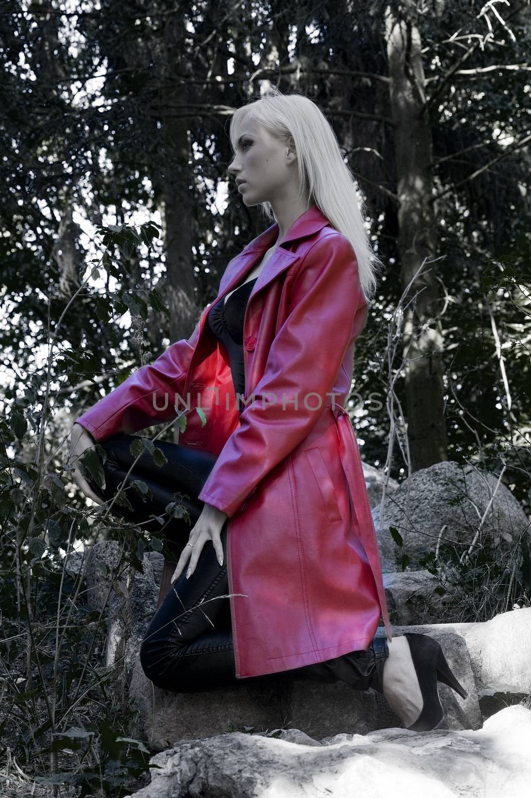 blond sexy woman outside in park in a fashion dark shot wearing a long red coat and sexy fitted black skin trousers