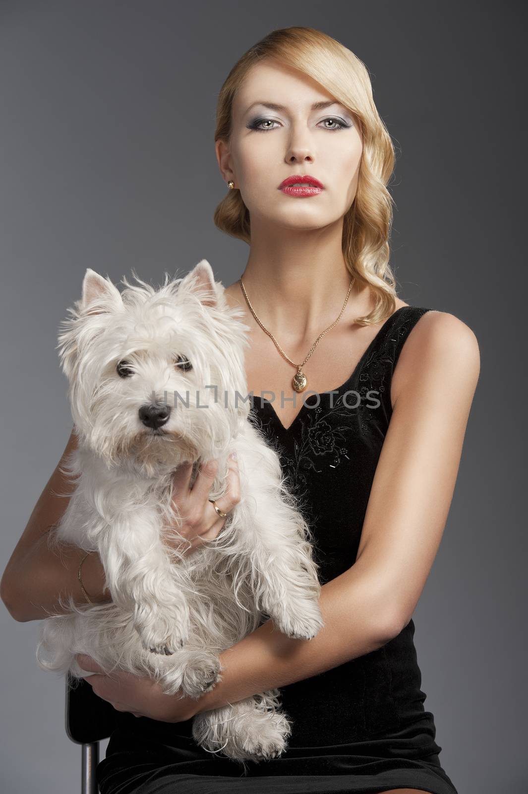 young elegant blond woman wearing black dress with an old fashion hairtyle and necklace jewellery, she is in front of the camera, takes a dog in her arms and looks in to the lens