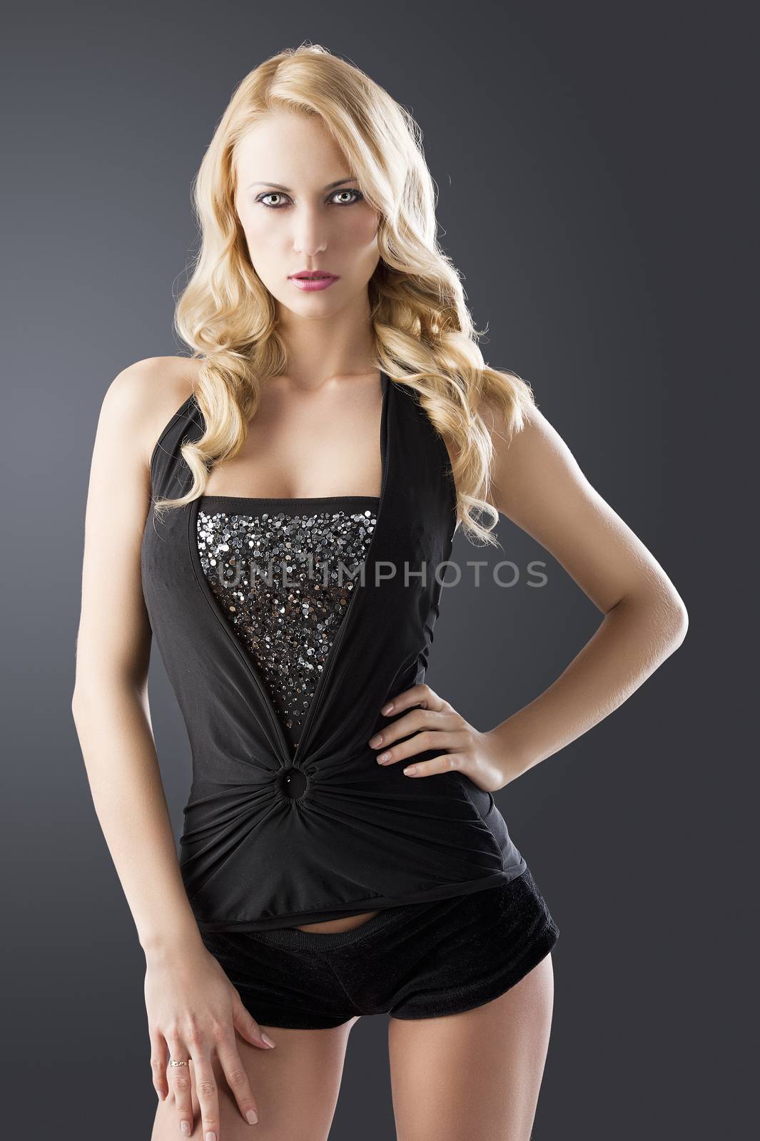elegant and sexy woman in black with shorts and with a curly hair style over black background, she is in front of the camera, looks in to the lens and her left hand is on the left hip