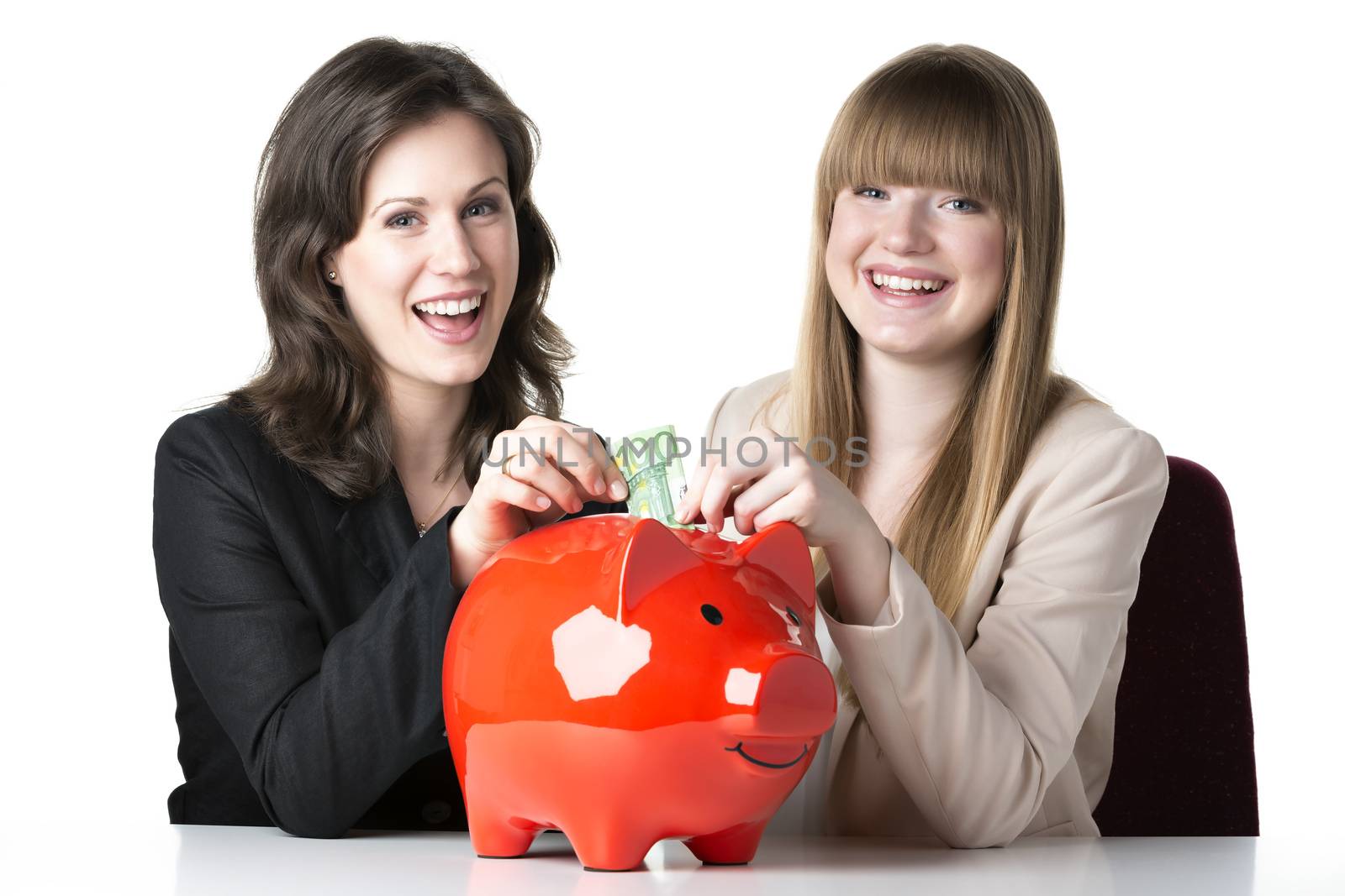 Two happy women sitting at a table and throwing 100 euro in a red piggy bank