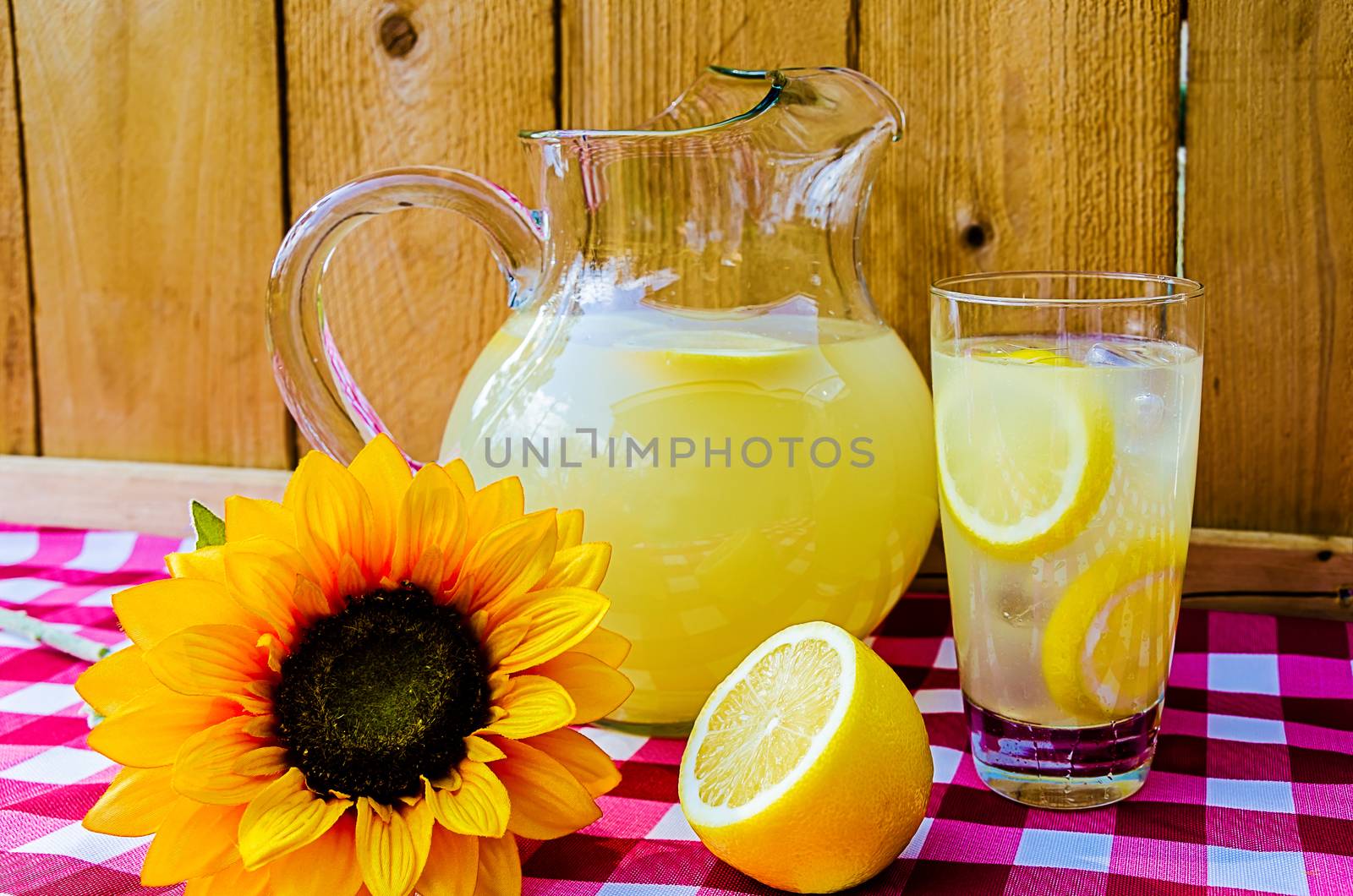 Lemonade with sliced lemons, pitcher, and sunflower on gingham table cloth.