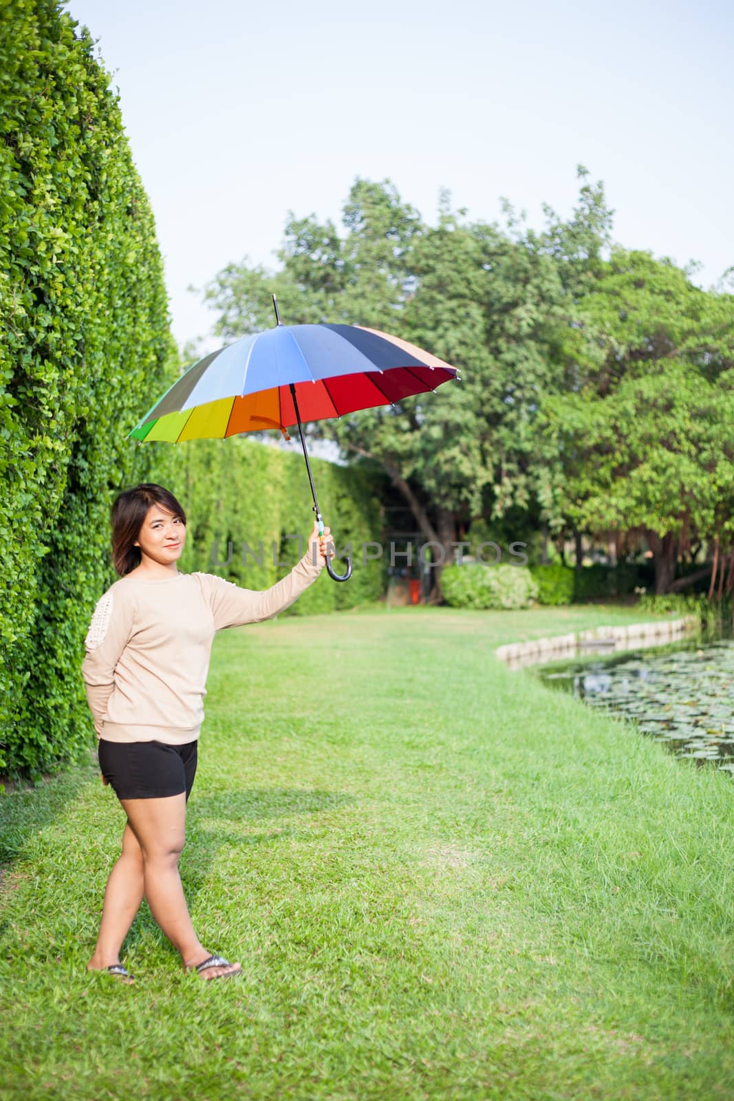 Asian woman holding an umbrella in the park. Stood on the lawn beside a tree.