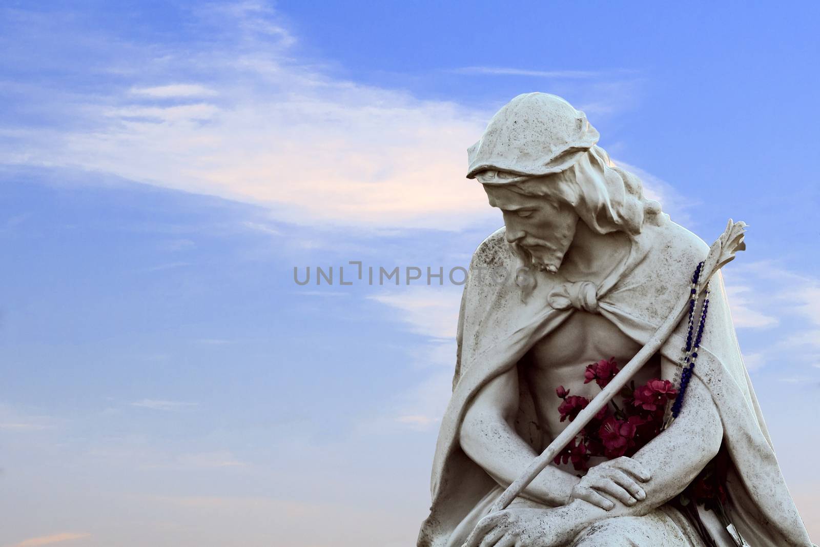 Sorrowful jesus statue with flowers on a blue sky with wispy clouds.