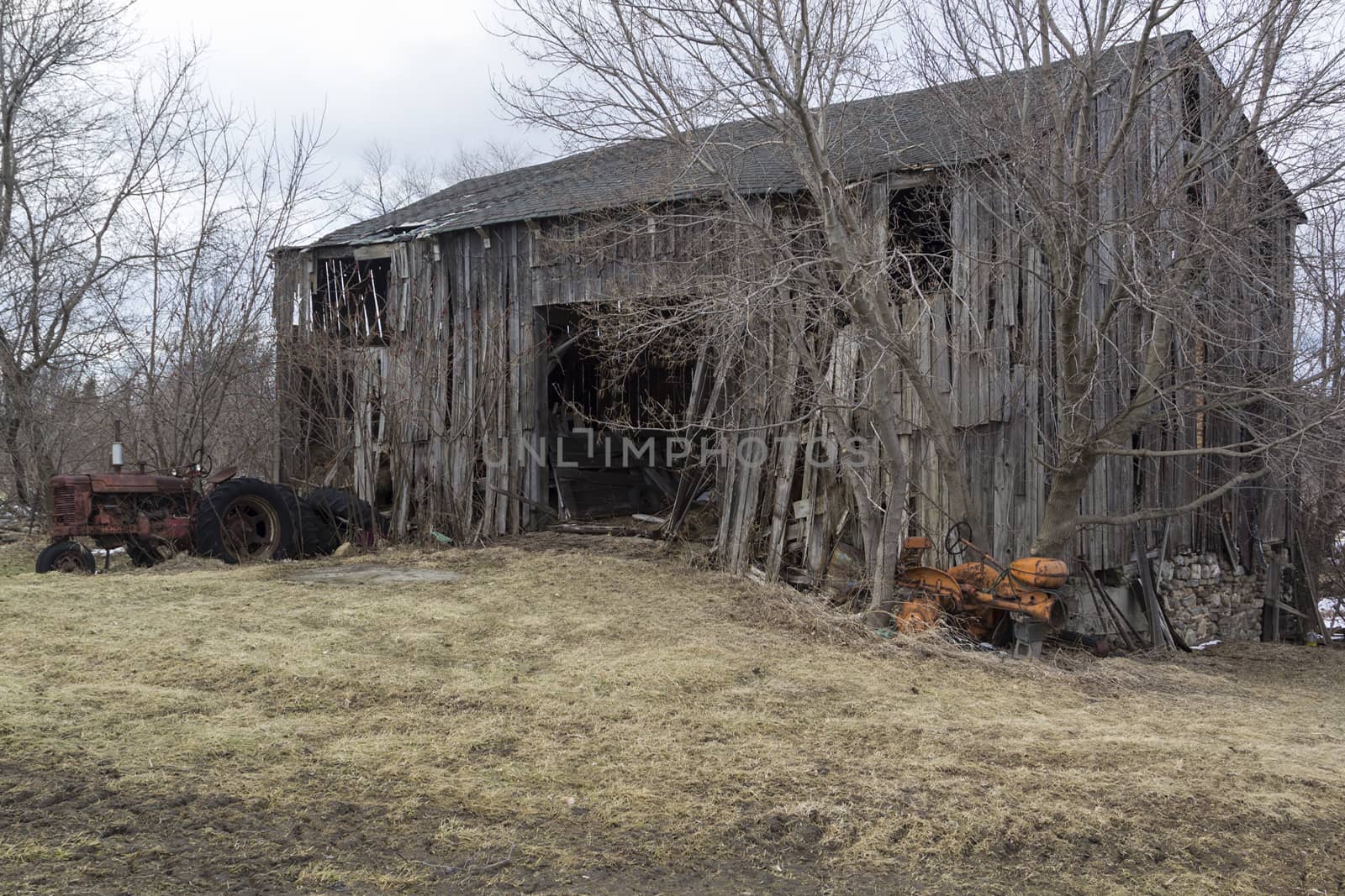 Old dilapidated and abandoned barn with rusty tractors and bare winter trees. 
