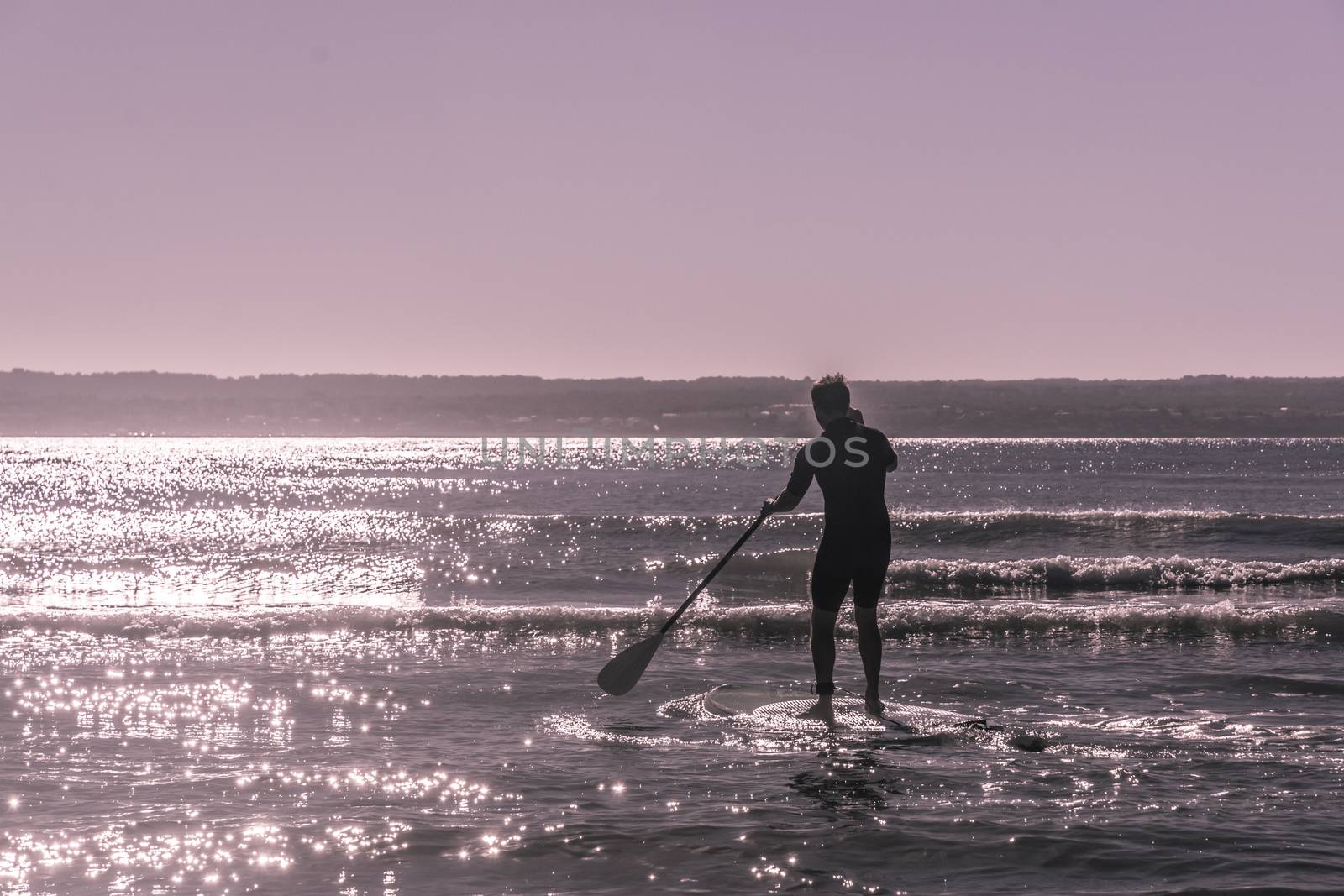 Young unrecognizable man paddle surfs the waves in Majorca, Balearic islands, Spain. Sunny violet filter treatment.
