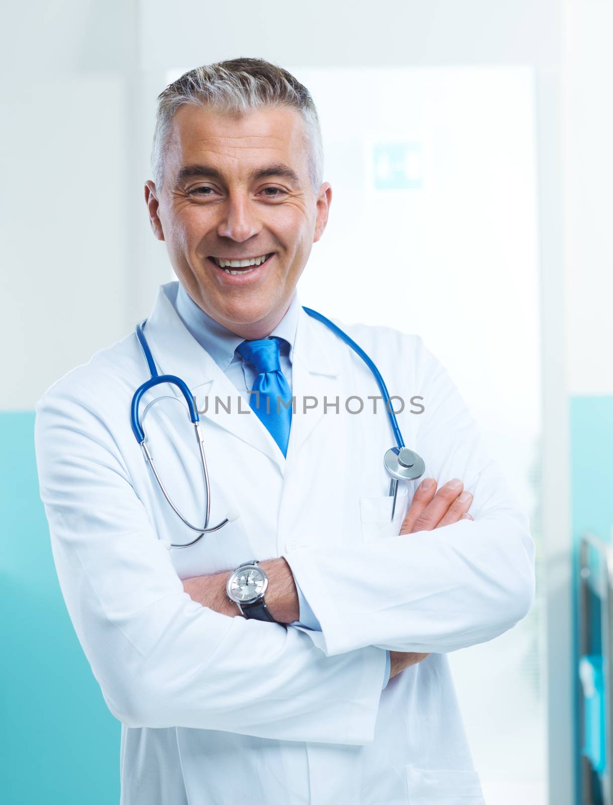 Friendly doctor portrait with arms crossed and medical equipment in teh background.