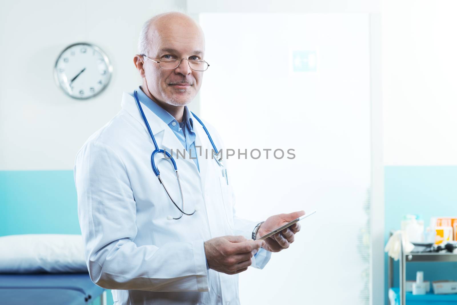 Senior doctor with tablet and medical equipment in the background.