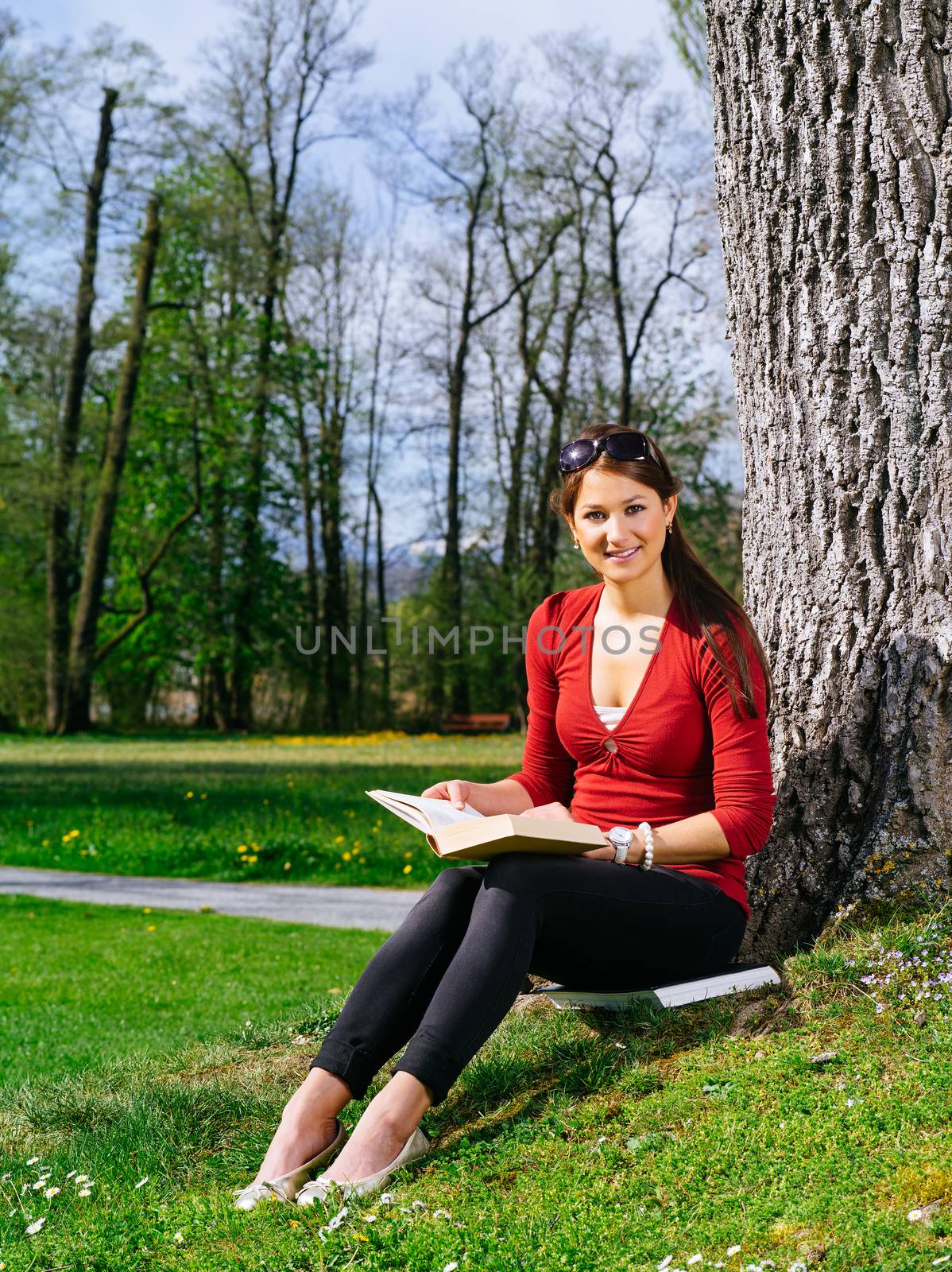 Photo of a beautiful young woman reading a book sitting against a tree in early spring.
