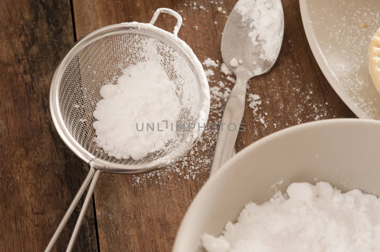 Kitchen sieve filled with icing sugar lying on a wooden kitchen counter alongside a mixing bowl while baking or cooking pastries and food