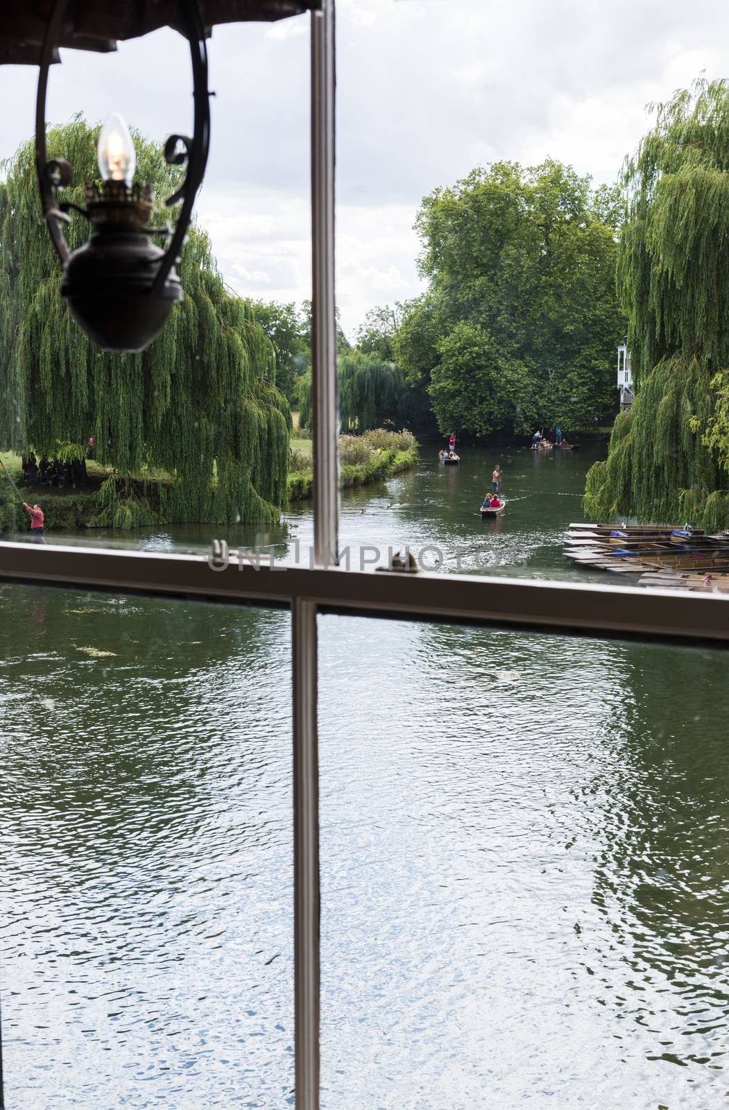 CAMBRIDGE, UK - AUGUST 18: River Cam with tourist punters in gondolas and park full of trees in the background viewed through old window with oil-lamp styled light. August 18, 2013 in Cambridge.
