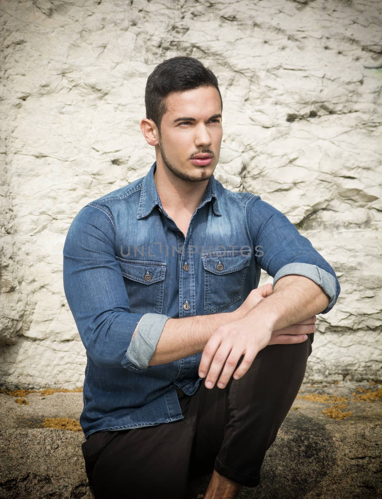 Handsome young man with denim shirt sitting outdoors in front of stone 